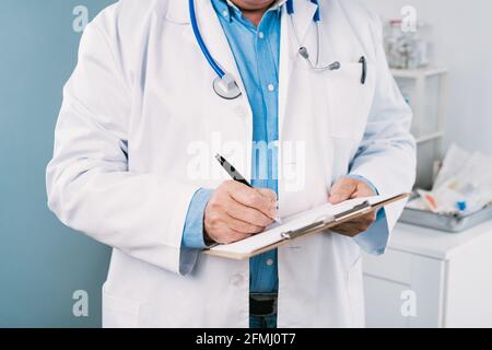 Crop anonimo medico maschile in accappatoio medico con stetoscopio e. appunti prendendo appunti su foglio cartaceo in ospedale Foto Stock