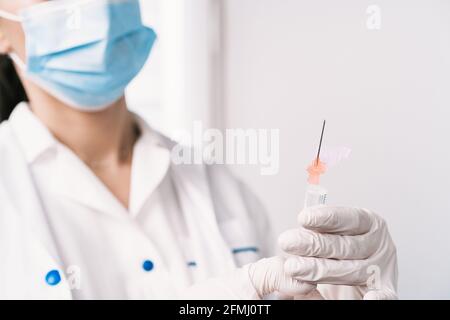 Medica femminile tagliata e non riconoscibile in maschera protettiva e con vaccino da coronavirus in siringa in piedi in sala medica luminosa di ospedale Foto Stock
