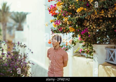 Giovane uomo sognante in piedi con le mani dietro vicino bianco rurale case decorate con fiori in vaso multicolore in fiore nel giorno di sole e guardando aw Foto Stock