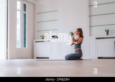 Vista laterale da terra della femmina sottile seduta sul tappetino In Vajrasana con le mani di preghiera e praticare yoga a casa Foto Stock