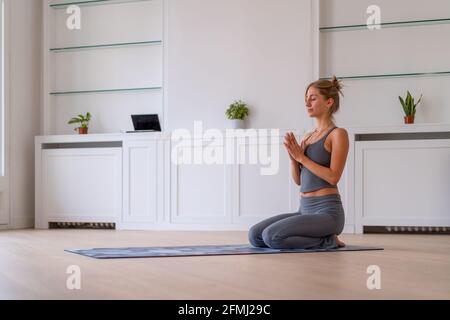 Vista laterale da terra della femmina sottile seduta sul tappetino In Vajrasana con le mani di preghiera e praticare yoga a casa Foto Stock