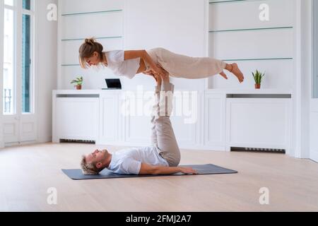 Vista laterale della fidanzata che solleva la ragazza mentre fa acro yoga insieme a casa e tenendo le mani Foto Stock