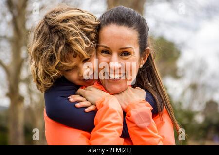 Ragazzo affascinante abbracciando madre sorridente mentre si siede in un legno panca che guarda via di giorno Foto Stock