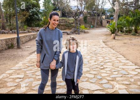 Madre in abbigliamento sportivo che tiene il ragazzo a mano mentre passeggi passerella e parlare contro gli alberi lookinga strada Foto Stock