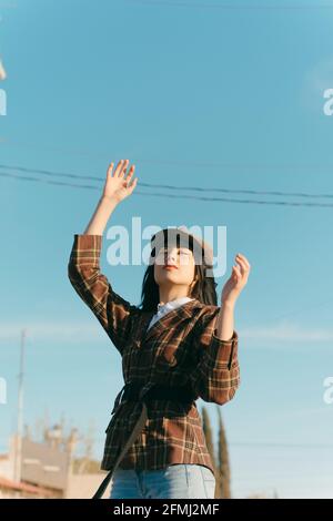 Giovane donna etnica sognante in beret e giacca a scacchi in piedi sotto il cielo nuvoloso e guardando in alto alla luce del sole Foto Stock