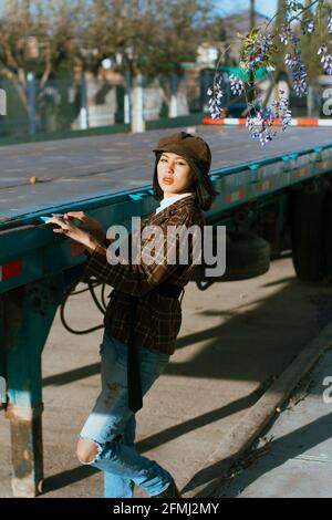 Vista laterale della giovane donna etnica elegante in beret contro rimorchio vecchio che guarda la fotocamera alla luce del sole Foto Stock