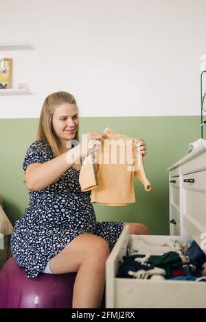 Donna allegra in attesa in abito ornamentale seduta su palla yoga con vestiti per bambini contro la commode in camera Foto Stock