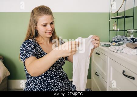 Donna allegra in attesa in abito ornamentale seduta su palla yoga con vestiti per bambini contro la commode in camera Foto Stock