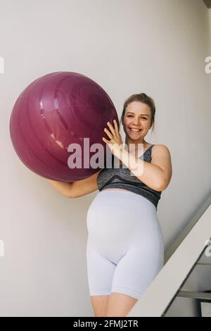 Giovane donna sorridente in abiti sportivi con palla yoga passeggiate su scale in casa Foto Stock