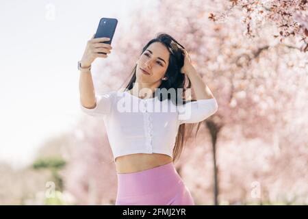 Giovane tenera femmina che prende selfie con il cellulare mentre tocca i capelli di luce diurna Foto Stock