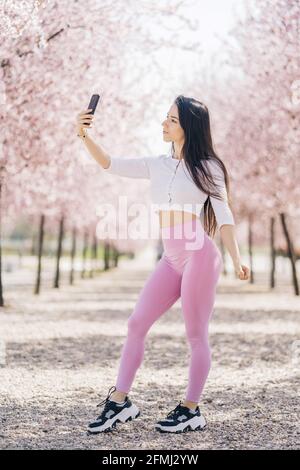 Giovane tenera femmina che prende selfie con il cellulare mentre si sta in piedi parcheggio Foto Stock