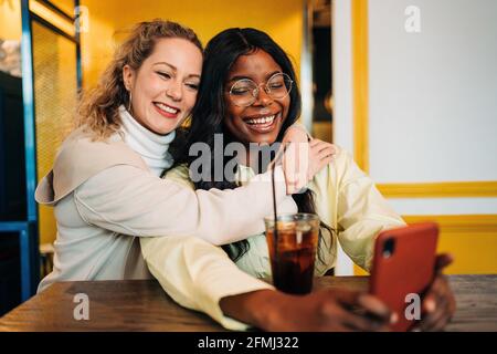 Felice multirazziale femmina migliori amici abbracciare in caffè e prendere scatta da solo sullo smartphone mentre ti godi il weekend insieme Foto Stock