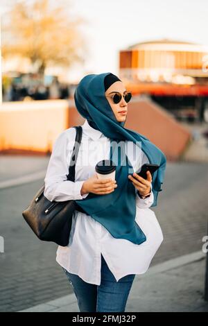 Donna etnica con sciarpa e occhiali da sole eleganti che camminano con take-away bere in strada e tenere in mano smartphone e guardare lontano Foto Stock
