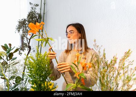 Giovane fiorista femminile concentrato in grembiule e occhiali arrangiando fragranti fiori d'arancio in vaso mentre si lavora in negozio di fiori Foto Stock