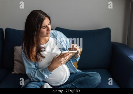 Donna incinta concentrata seduta sul divano e scrittura in blocco note mentre si rilassa in un accogliente soggiorno Foto Stock
