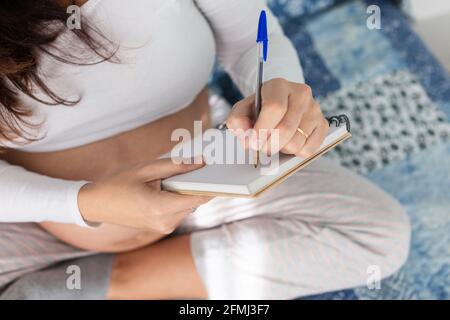 Dall'alto della donna incinta seduta sul letto e la scrittura in taccuino di mattina Foto Stock