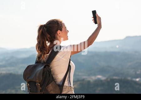 Vista posteriore ragazza hiker felice con zaino che prende selfie sullo smartphone mentre ci si trova in cima a una collina verdeggiante Foto Stock