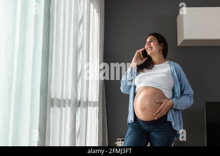 Sorridente donna incinta che tocca il ventre mentre si è in piedi nella stanza a. telefono cellulare a casa e in conversazione Foto Stock