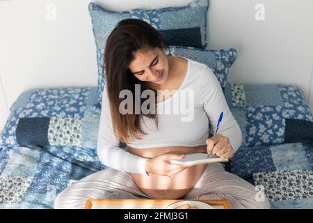 Dall'alto della donna incinta seduta sul letto e la scrittura in notebook durante la colazione al mattino Foto Stock