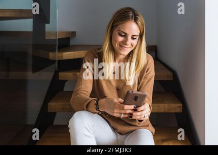 Felice donna seduta sulle scale a casa e navigando smartphone Foto Stock