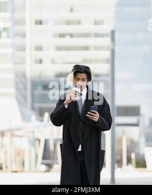 Giovane uomo etnico ben vestito executive con cellulare e caldo drink da andare in città Foto Stock