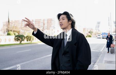 Giovane elegante imprenditore etnico maschile con mano in tasca dimostrare gesto di vittoria mentre si guarda lontano contro la strada della città Foto Stock