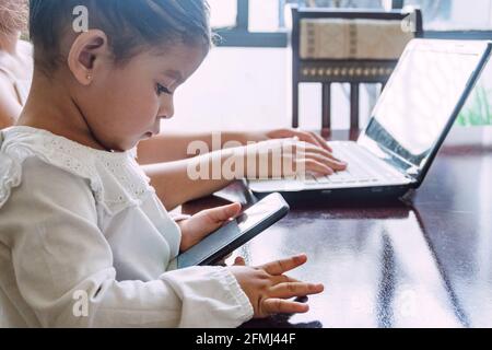 Vista laterale di etnico bambina che naviga telefono cellulare mentre seduto al tavolo con la madre di raccolto che scrive sul laptop a. casa Foto Stock