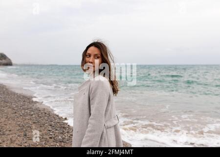 Affascinante donna tranquilla in mantello in piedi sulla spiaggia di sabbia e. guardando via Foto Stock