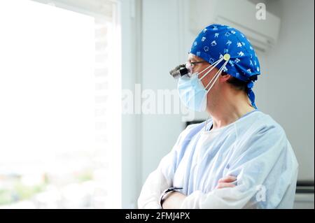 Medico maschile anonimo in uniforme e maschera sterile guardando via con braccia ripiegate contro la finestra in ospedale Foto Stock