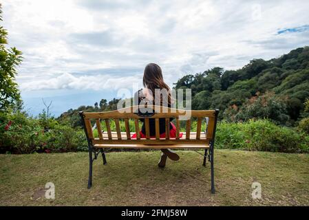 Vista posteriore di anonimo turista femminile seduta con gambe incrociate Su panchina contro alberi lussureggianti sotto il cielo nuvoloso in Thailandia Foto Stock