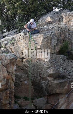 Arrampicatore maschio in casco protettivo seduto su roccia e fissaggio imbracatura prima della discesa dalla montagna Foto Stock