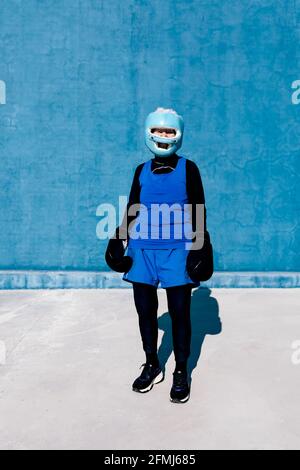 Femmina maturo di lunghezza intera in abbigliamento sportivo e guanti da boxe in piedi con casco contro parete blu e guardando la fotocamera Foto Stock
