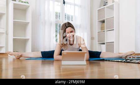 Sorridente donna flessibile che pratica yoga a Upavista Konasana mentre si siede su mat e guardare video tutorial online su tablet all'indirizzo casa Foto Stock