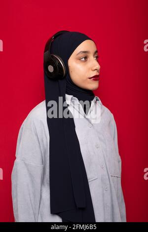 Attraente donna musulmana in abiti casual e hijab ascolto musica preferita tramite cuffie su sfondo rosso in studio Foto Stock