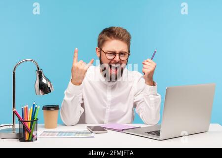 Un lavoratore in ufficio positivo e eccitato che mostra movimenti rock e roll sedersi sul posto di lavoro con il blocco note e la matita del portatile finito con successo repo annuale Foto Stock