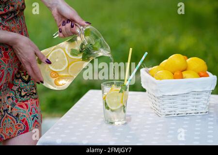 Le mani femminili versano una rinfrescante limonata fredda con i limoni nel ghiaccio e la menta da una caraffa in un bicchiere su un tavolo da giardino estivo su un picnic. Soft selective fo Foto Stock