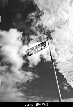 La bandiera dell'Unione britannica (Union Jack) su un palo di bandiera contro un cielo blu con soffici nuvole bianche. Fotografia in bianco e nero. Foto Stock