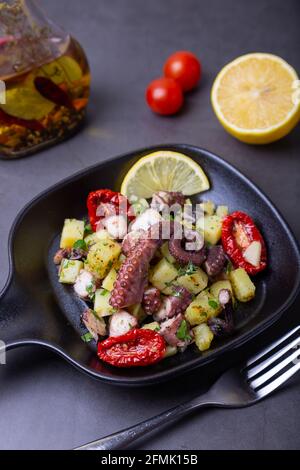 Insalata tiepida con polpo, patate, pomodori secchi, aglio e limone in una piccola padella nera. Primo piano, sfondo nero. Foto Stock