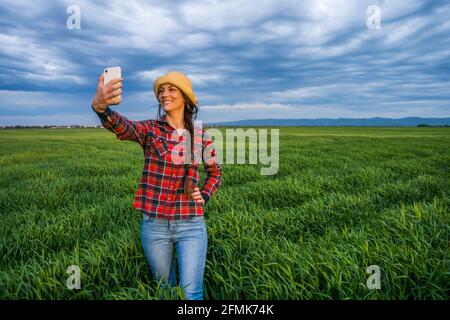 Orgogliosa agricoltore femminile è in piedi nel suo campo di orzo e godendo il tramonto. Sta prendendo selfie. Foto Stock