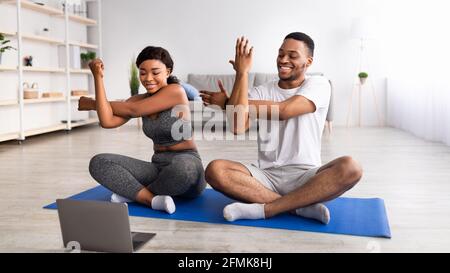 Giovane coppia nera che pratica yoga o pilates, seduto in posa lotus, allungando le braccia di fronte al computer portatile durante l'allenamento domestico, panorama. SpO Foto Stock