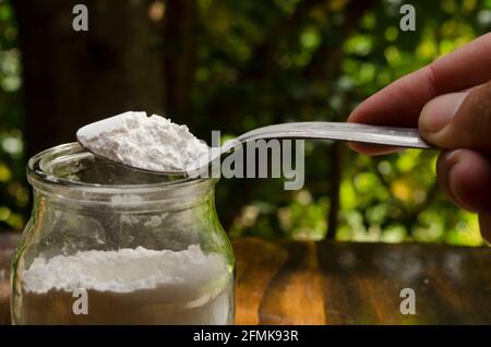 polvere da forno prelevata da un vasetto di vetro in cucchiaino da tè su tavolo di legno con sfondo naturale Foto Stock