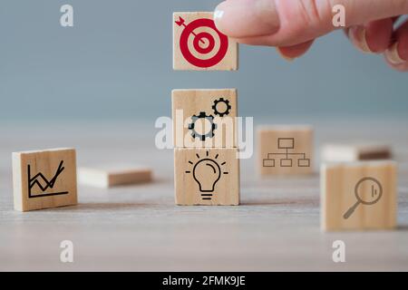 Strategia aziendale, piano d'azione, concetto di gestione. Mano di uomo d'affari che mette il blocco di cubo di legno con l'icona di bersaglio sopra altri cubi Foto Stock