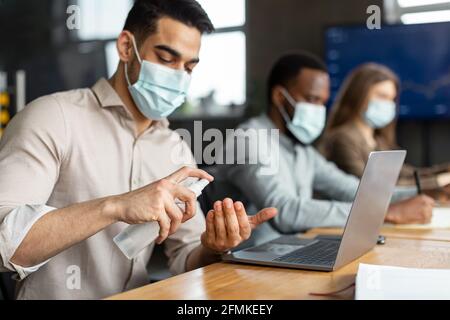 Misure preventive, arrestare la diffusione del coronavirus. Ritratto di giovane uomo arabo manager spray disinfettante a portata di mano, fuoco selettivo. Diversi dipendenti weari Foto Stock