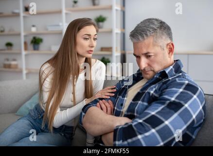 Concetto di argomento familiare. Infelice uomo maturo seduto sul divano, sua moglie lo consolava, cercando di fare pace dopo il conflitto a casa. Sposato caucasico Foto Stock