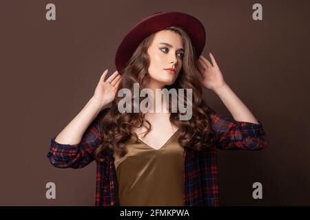 Donna elegante in camicia plaid e cappello fedora che si posano sopra sfondo marrone Foto Stock