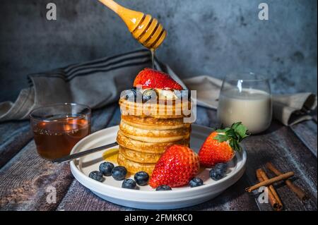 Una pila di frittelle soffici e soffici con sciroppo d'acero versato da bastone, mirtillo fresco, fragola e un bicchiere di latte su tavola di legno, delizioso essiccatoi Foto Stock