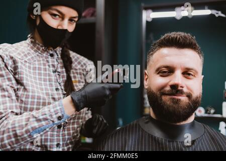 Soddisfatto giovane cliente maschio di barbershop si siede in poltrona ottiene il taglio di capelli da giovane donna parrucchiere indossando maschera protettiva e guanti. Foto Stock