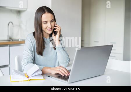 Giovane donna di charme freelancer sta parlando su smartphone lavorando con il laptop da casa, la donna ha una piacevole conversazione telefonica lavorando a distanza, connettersi con i clienti tramite telefono cellulare Foto Stock