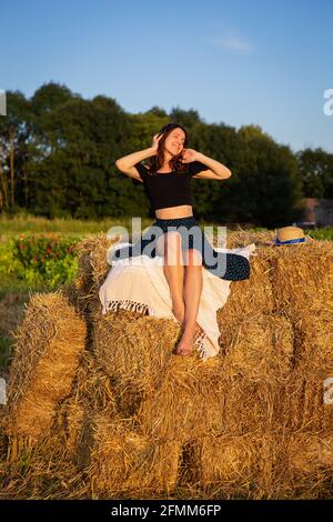 Giovane bella donna guarda il sole che tramonta, si siede su un grande mucchio di balle di paglia Foto Stock