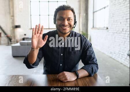 Un uomo indovino cordiale e disponibile che indossa un dispositivo auricolare e una camicia casual intelligente guarda la fotocamera e il saluto interlocutore, rappresentante di supporto in contatto, indù ragazzo in cuffie coinvolti riunione Foto Stock